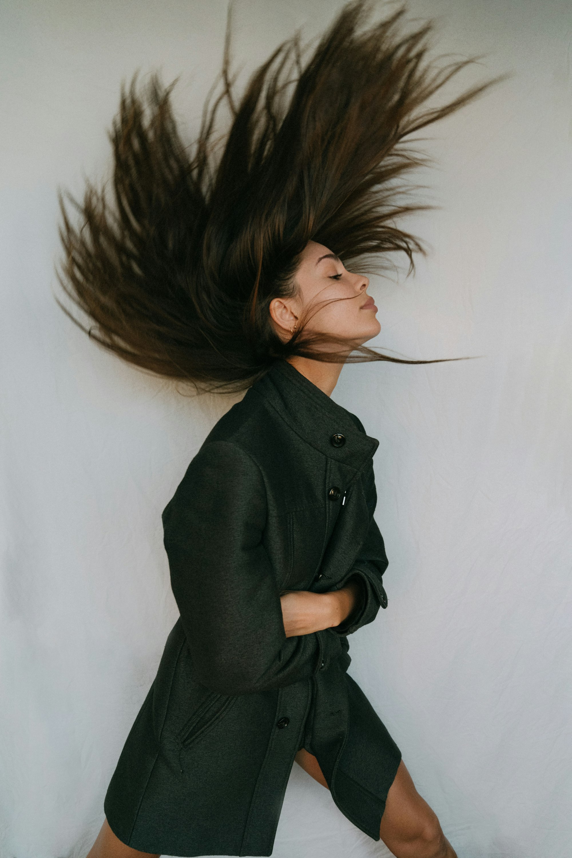 Woman in a dark coat tossing long brunette hair upwards against a pale wall