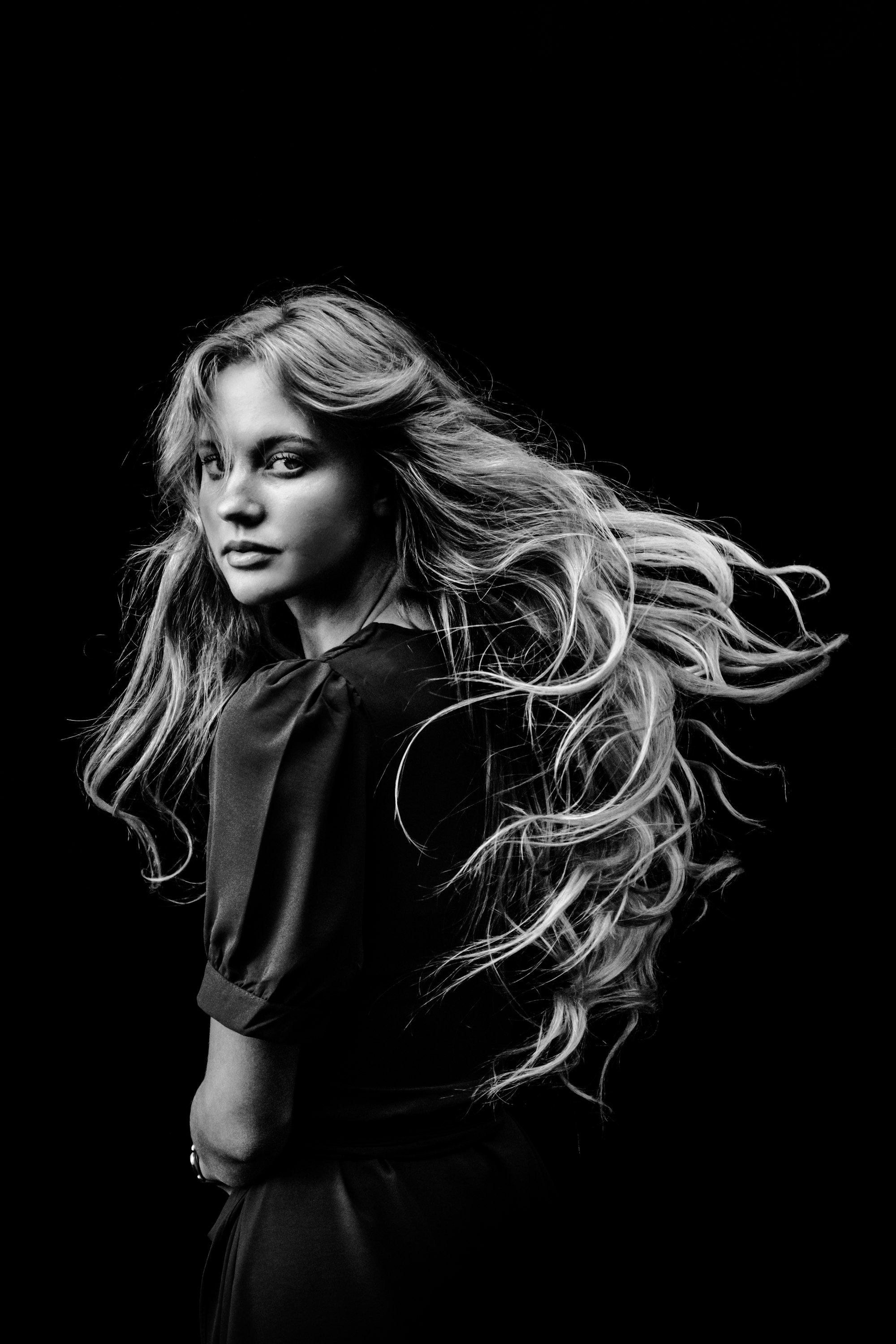 Black and white portrait of a woman glancing over her shoulder as her long, wavy hair sweeps through the air